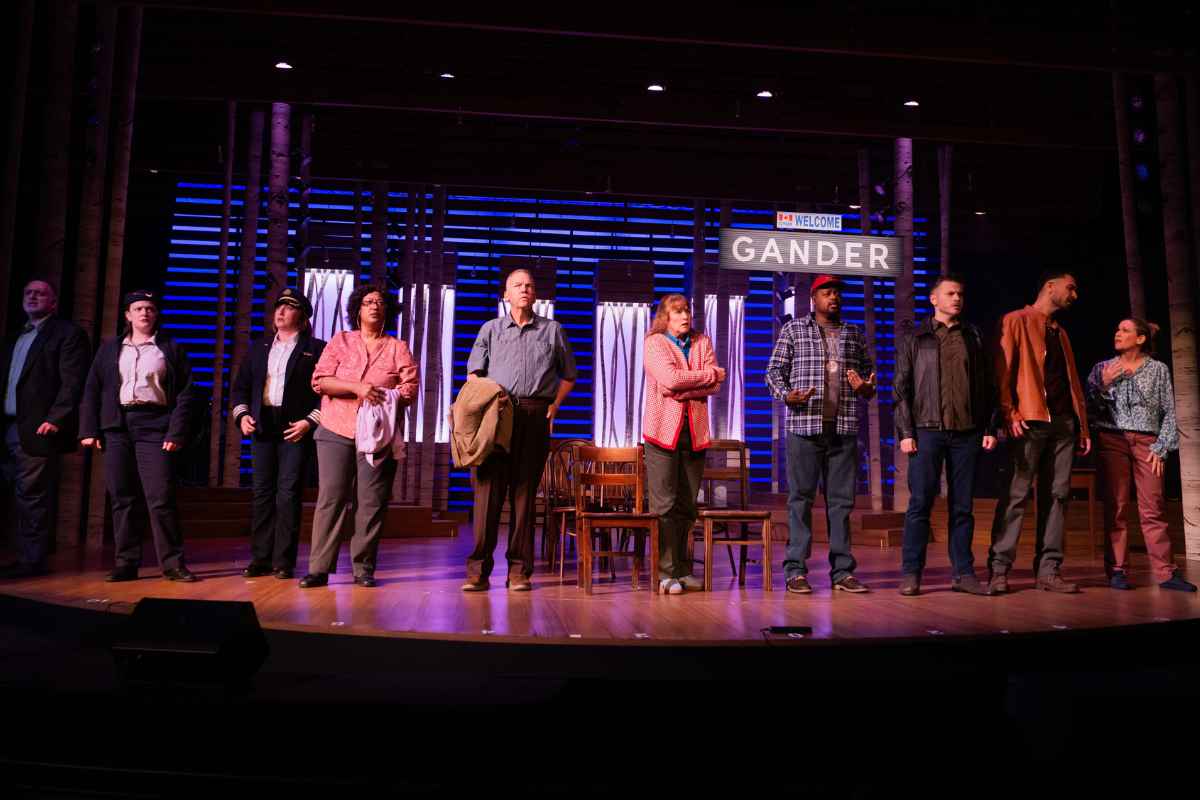 Group of ten actors on a stage standing in a line with concerned looks on faces. Sign over head reads Welcome Gander.